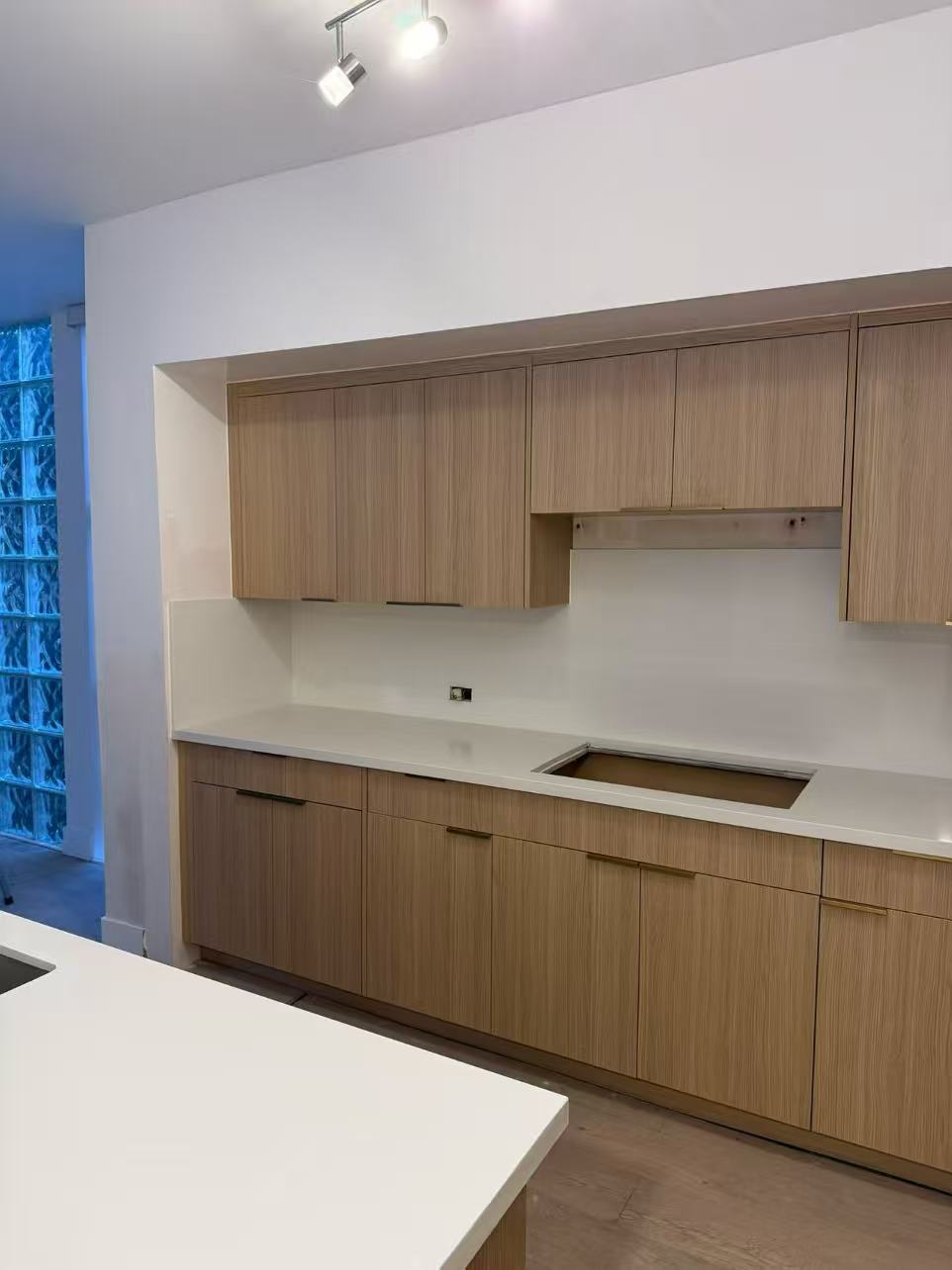 Light-oak frameless cabinets with white quartz countertops and glass-block window.